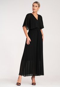 Czarna maxi sukienka z plisami, wykończona dekoltem w serek i lekko zwiewnymi rękawami-kapami. Wykonana z lekkiego materiału. Stylizowana z czarnymi sandałami na paski.