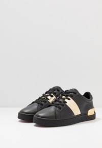 Par de zapatillas negras de corte bajo con cordones negros y paneles laterales dorados metálicos y acentos en el talón sobre una superficie blanca.