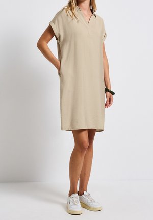 Kobieta w beżowej sukience do kolan z krótkim rękawem i kołnierzykiem oraz białych trampkach, stojąca z jedną ręką w kieszeni na białym tle.