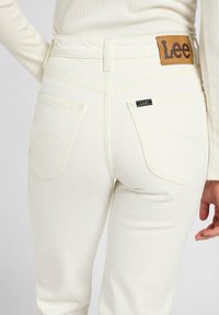 Pantalones vaqueros de mezclilla blanca con un acabado texturizado, que cuentan con bolsillos traseros y un parche de cuero marrón con el logo de Lee en la cinturilla.