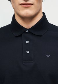 Κοντινό πλάνο ενός ατόμου που φοράει μαύρο πόλο μπλουζάκι με γιακά, τρία κουμπιά και ένα μικρό κεντητό λογότυπο στο στήθος.