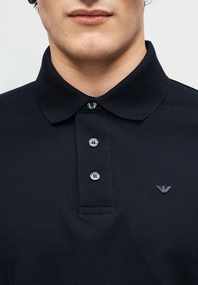 Κοντινό πλάνο ενός ατόμου που φοράει μαύρο πόλο μπλουζάκι με γιακά, τρία κουμπιά και ένα μικρό κεντητό λογότυπο στο στήθος.