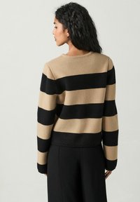 Sweter dziany z szerokimi poziomymi paskami w kolorze czarnym i beżowym, okrągłym dekoltem, ściągaczami przy rękawach oraz luźnym krojem, w połączeniu z czarnymi spodniami.