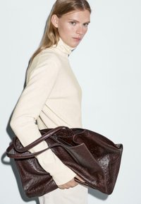 Brązowa skórzana torba tote o teksturowanej powierzchni z dwiema długimi rączkami, trzymana przez osobę noszącą kremowy sweter z golfem.