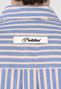 Dos du cou et des épaules d'une personne portant une chemise rayée bleue et beige avec un patch blanc marqué « Interdit » en noir.