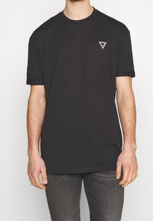 Homme portant un t-shirt noir uni avec un petit logo géométrique blanc sur le côté gauche de la poitrine, associé à un jean gris foncé, debout devant un fond blanc.