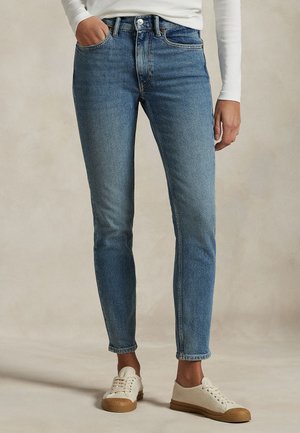 Une personne porte un jean bleu à jambes droites, un haut blanc et des baskets couleur crème, une main dans une poche avant.