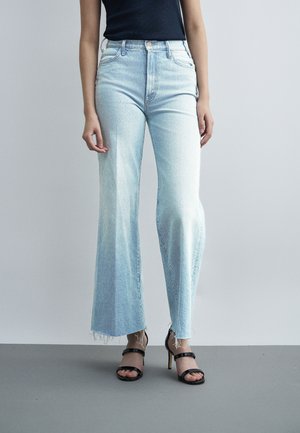 Žena v svetlomodrých džínsach s vysokým pásom a rozšírenými nohavicami, s neupravenými lemy, a čiernych sandáloch na vysokom podpätku s remienkami, stojaca na jednofarebnom pozadí.