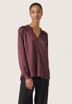 Burgundy long-sleeve blouse with a V-neck, pleated details, and button cuffs. Smooth texture, slightly loose fit, and side slits at the hem.