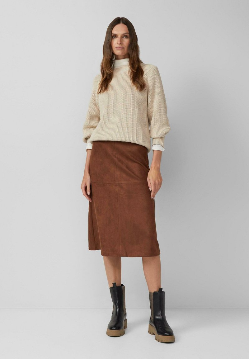 Femme portant un pull en tricot beige, une jupe midi en daim marron et des bottes noires à semelles épaisses, sur fond neutre.