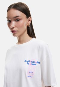 Weißes, übergroßes T-Shirt aus Baumwolle. Mit mehrfarbigem Text und Grafik auf der Vorderseite, mit blauen und pinken Akzenten. Rundhalsausschnitt.