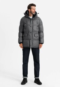 Giacca a vento grigia con cappuccio, caratterizzata da dettagli con bottoni e quattro tasche frontali. Indossata sopra jeans scuri e stivali neri, con una texture liscia.