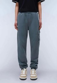 Graue Sweatpants in einer lockeren Passform, mit einem elastischen Bund und Seitentasche. Kombiniert mit beigen und weißen Turnschuhen.
