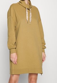 Khaki klänning med huva och långa ärmar, ribbade ärmslut och snörning vid halsen. Har sidofickor med dragkedja vid fållen för justerbar passform.