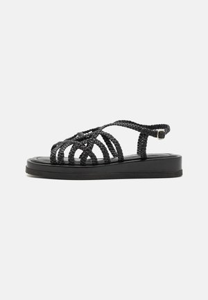 Sort flad sandal med flettede stropper og ankelspænde på en ensartet lys baggrund.