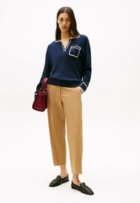 Un pull marin avec un col blanc et une poche, des bordures crème, un pantalon beige et des mocassins noirs. Tient un petit sac à main bordeaux.