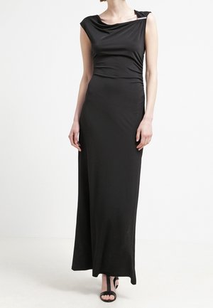 Mujer vistiendo un vestido negro sin mangas, largo hasta el suelo, con escote drapeado y tacones abiertos negros, de pie sobre un fondo blanco.