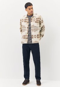 Chemise beige clair à boutons avec des motifs géométriques en tons bruns et rouille, portée avec un jean bleu foncé et des chaussures marron.