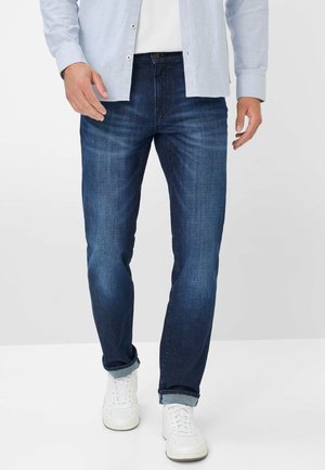 Dark blue denim jeans with a straight cut, featuring a subtle texture and slight fading. Worn with rolled cuffs and white sneakers.