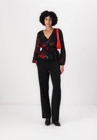 Blusa nera a fiori con motivi di rose rosse e verdi, design peplum e maniche lunghe; abbinata a pantaloni neri sartoriali e una borsa rossa.