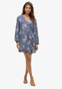 Vestido envolvente azul marino con patrón de puntos blancos, mangas largas abullonadas, dobladillo acampanado y bordes con volantes. Modelo lleva sandalias de tacón cuadrado en color beige.