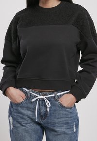 Svart kort sweatshirt med texturerad spetsdetalj vid axlarna, kombinerad med blå slitna jeans och en vit dragsko.