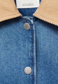 Denim jacket with a beige corduroy collar, featuring silver snap buttons. The fabric has a textured weave with blue hues and visible stitching.