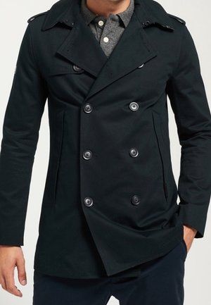 Mężczyzna ubrany w ciemny płaszcz dwurzędowy narzucony na szarą koszulę zapiętą na guziki, z rękami w kieszeniach, stojący na białym tle.