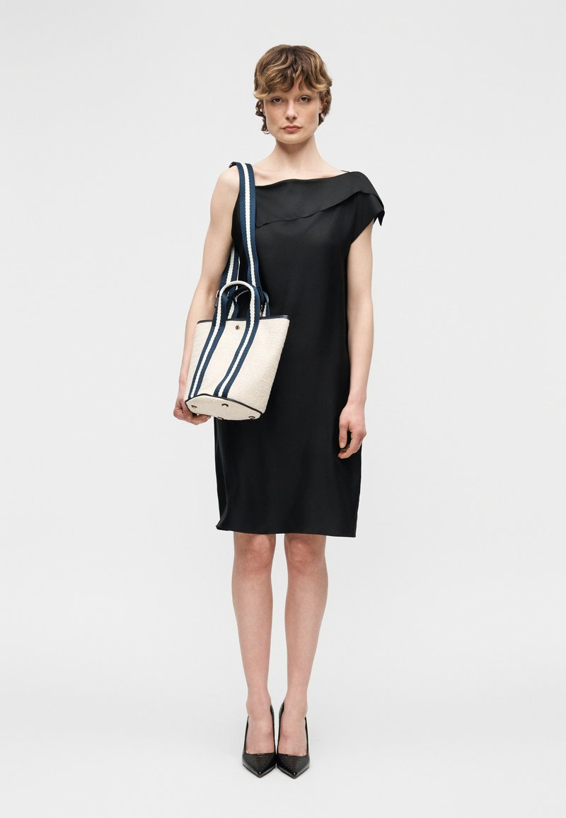 Γυναίκα με μαύρο αμάνικο φόρεμα και μαύρες γόβες, κρατώντας μια λευκή τσάντα ώμου με λαβές με ναυτικό μπλε ρίγες στον ώμο της.