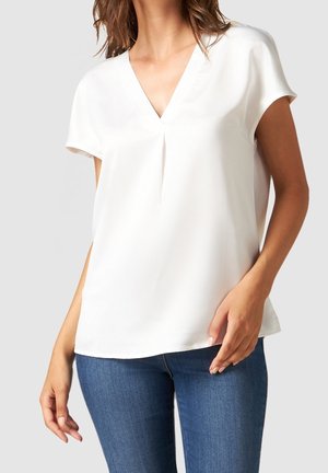 Frau trägt eine weiße V-Ausschnitt-Bluse mit kurzen Ärmeln, kombiniert mit blauen Jeansstoff-Jeans vor einem schlichten Hintergrund.