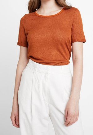 Femme portant un haut orange brûlé à manches courtes, rentré dans un pantalon blanc taille haute, debout devant un fond uni.