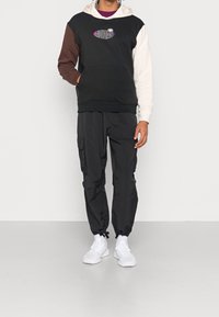 Person som bär en svart Adidas-hetja med färgblockerade ärmar, svarta cargobyxor, vita strumpor och vita sneakers som står mot en plain vägg.