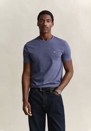 Homme portant un t-shirt bleu ajusté à manches courtes, un jean foncé, une ceinture noire et une montre-bracelet argentée, debout avec une main dans la poche.