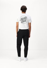 Witte t-shirt met een zwart "Santa Cruz" logo op de achterkant, gecombineerd met zwarte tapered broek en witte sneakers. Eenvoudig en modern ontwerp.