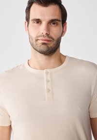 Beige kortermet T-skjorte laget av myk stoff, med rund hals og detalj med tre knapper foran.