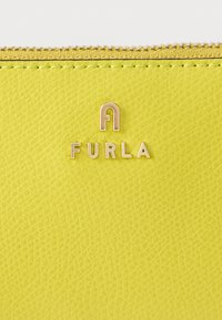 Κίτρινη επιφάνεια από δέρμα με υφή, με χρυσό λογότυπο Furla και φερμουάρ κατά μήκος της πάνω άκρης.