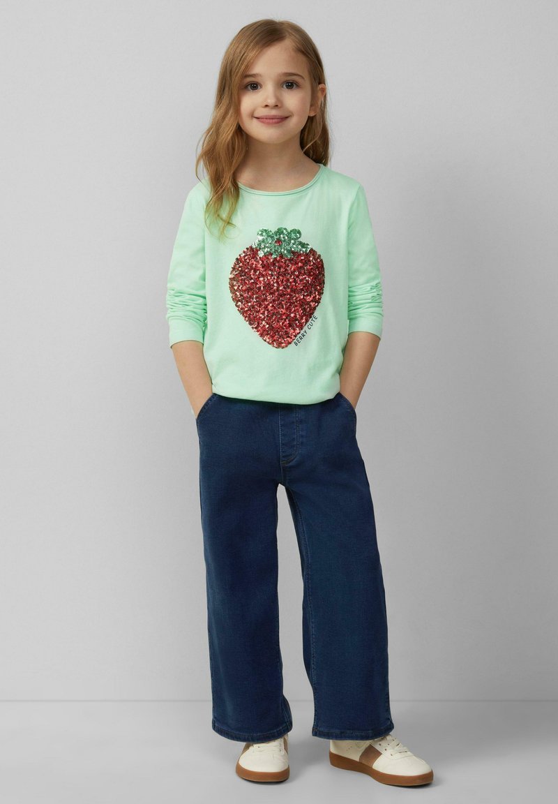 Mintgroene longsleeve shirt met een grote rode sequins aardbei graphic, gecombineerd met marineblauwe wide-leg jeans en witte sneakers.