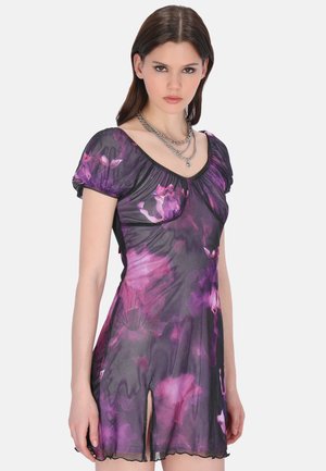 Jeune femme portant une robe fleurie violette transparente à manches courtes avec une fente à l'avant et des colliers en chaînes argentées superposés, debout devant un fond uni.