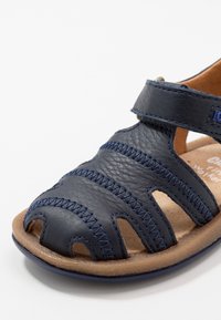 Mörkblå lädersandal med öppen design, detaljerad sömnad och en brun texturerad sula. Justerbar rem för en säker passform.