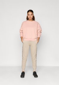 Világos rózsaszín pulóver  ribbelt ujjal, bézs nadrág oldalsó csíkokkal és fekete sneakers, ami egy laza, összerakott öltözetet alkot.