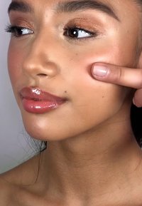 Close-up of a face with light brown skin, glossy lips, and soft makeup. A finger points to the cheek, highlighting its smooth texture.