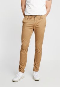 Chinos en coton beige avec une coupe slim, dotés d'une fermeture à bouton, de passants de ceinture et de poches latérales, associés à des baskets blanches.