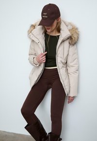 Giacca puffer beige con cappuccio bordato di pelliccia, top verde scuro, leggings marroni e stivali marroni fino al ginocchio. Cappello con il testo "FIDATI DEL PROCESSO".