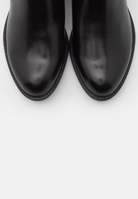 Botas de cuero negro hasta el tobillo con un acabado suave, punta redondeada y un detalle de costura mínima alrededor del borde. La suela está ligeramente elevada.