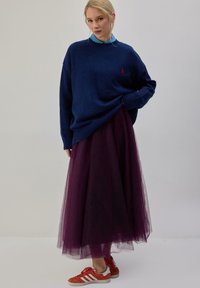 Maglione oversize blu navy con polsini a coste, indossato sopra una gonna in tulle viola, abbinato a sneakers rosse e bianche.