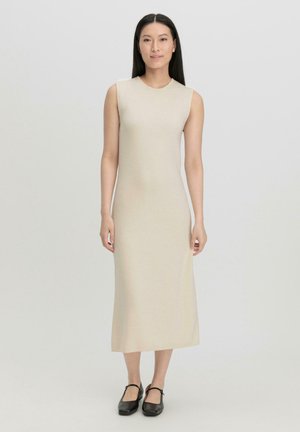 Abito beige senza maniche, lungo fino al ginocchio, realizzato in un tessuto liscio e elasticizzato con una silhouette aderente. Il modello indossa scarpe nere Mary Jane.
