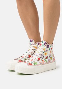 Wysokie sneakerzy wykonane z białego płótna z kolorowymi kwiatowymi i owocowymi wzorami, białymi sznurówkami oraz teksturowanymi gumowymi podeszwami.