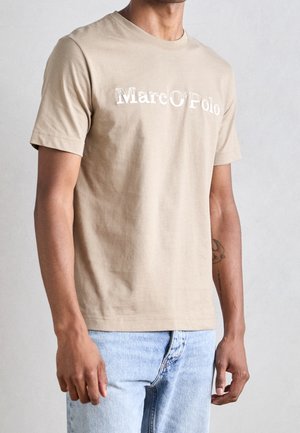 Beżowy bawełniany t-shirt z okrągłym dekoltem, krótkimi rękawami i logo "Marc O'Polo" w białym kolorze, zestawiony z jasnoniebieskimi jeansami.