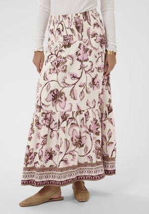 Kvinna bär en lång blommig kjol med rosa och bruna mönster, kombinerad med flätade platta skor och en vit långärmad topp med ribbad struktur.