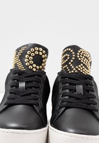 Sneaker in pelle nera con suola bianca. Presenta dettagli in oro a borchie sulla parte superiore nera. Include lacci neri e una superficie liscia.